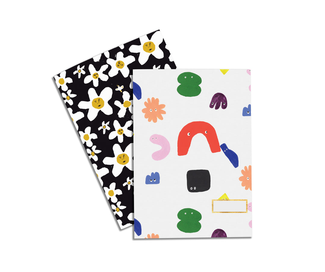 Illustrated Notebook Set by Atelier Mave with one design being a variety of shapes with googly eyes and funny faces and the other design being a floral pattern where the center of the flower includes funny faces. 