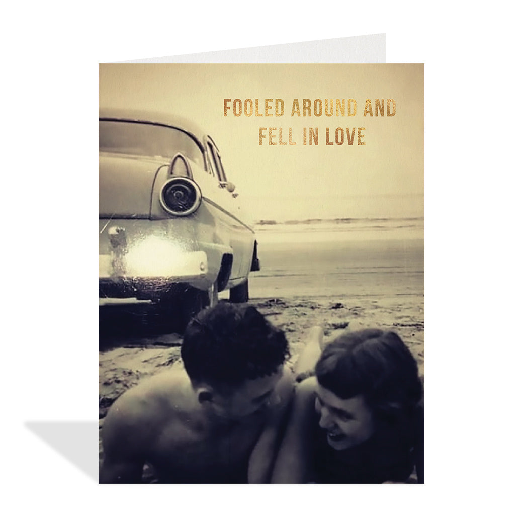 Black and white, vintage photographic card of a young couple laying down together on a beach giggling in front of an old car. Gold foil typography that reads "Fooled around and fell in love."