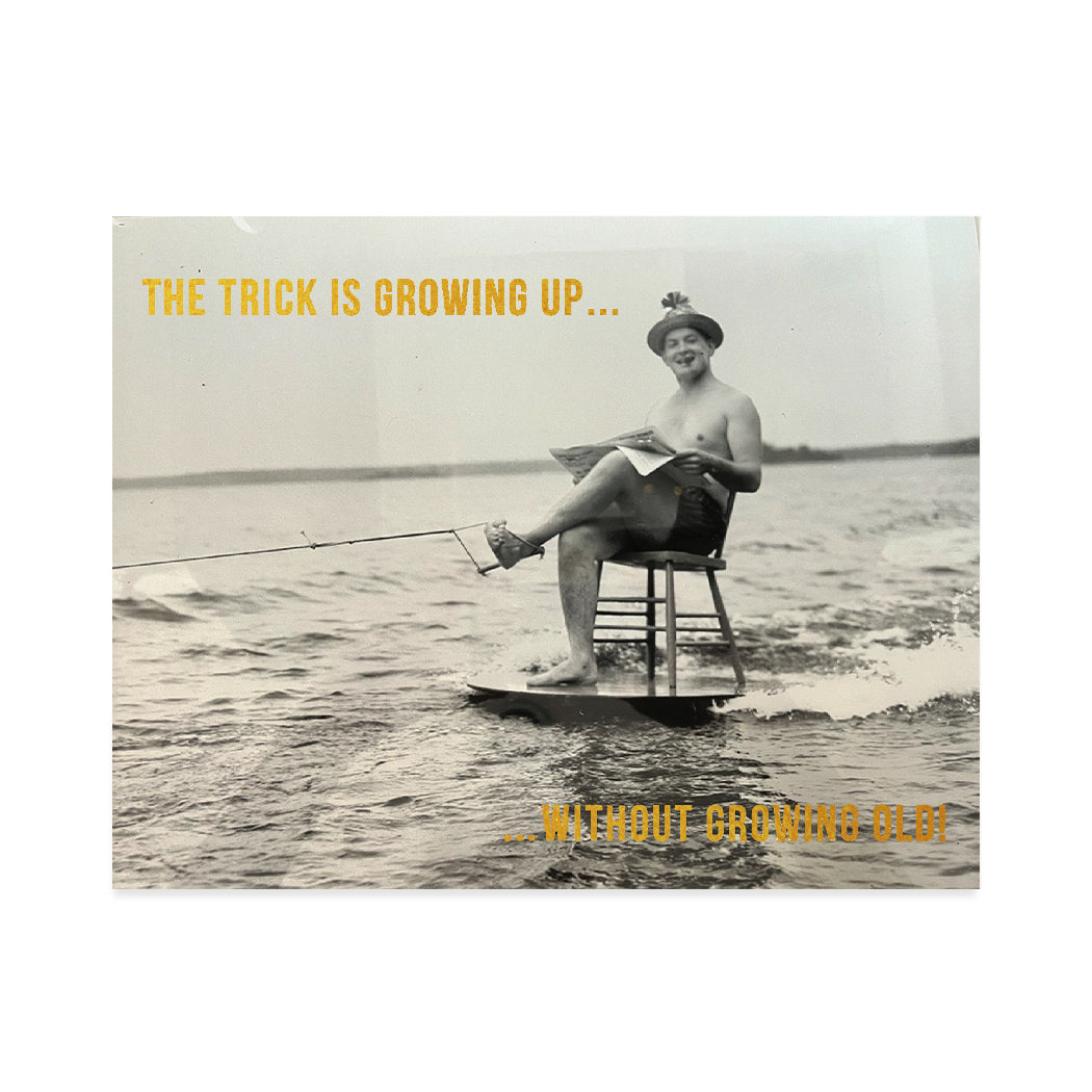 Vintage, black and white photographic card of a man reading the newspaper while sitting on a chair on a surf board riding the waves. Gold foil typography that reads "The trick is growing up without getting old".