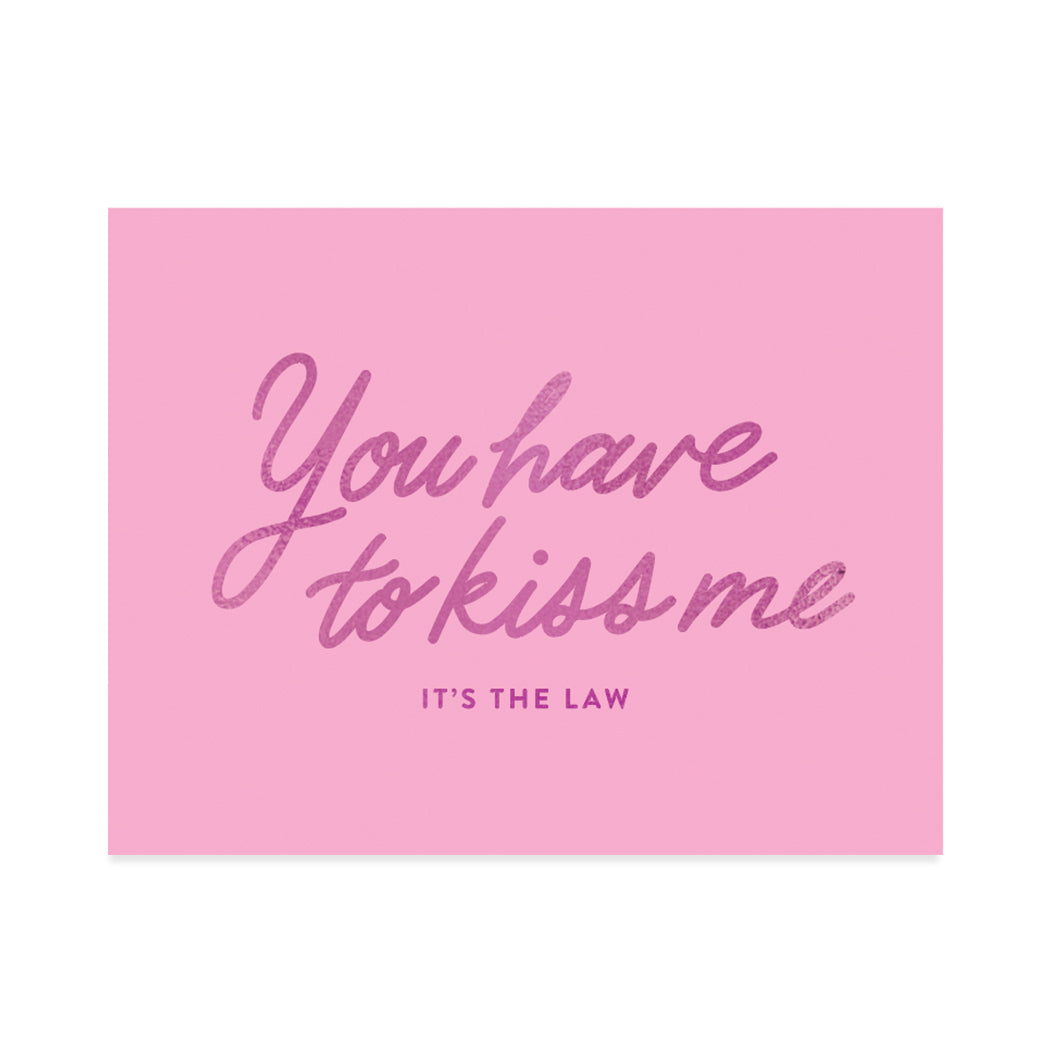 Greeting card designed by Halfpenny Postage. A simple design with a wild pink foil quote that reads, "you have to kiss me, it's the law" on a soft pink background. 