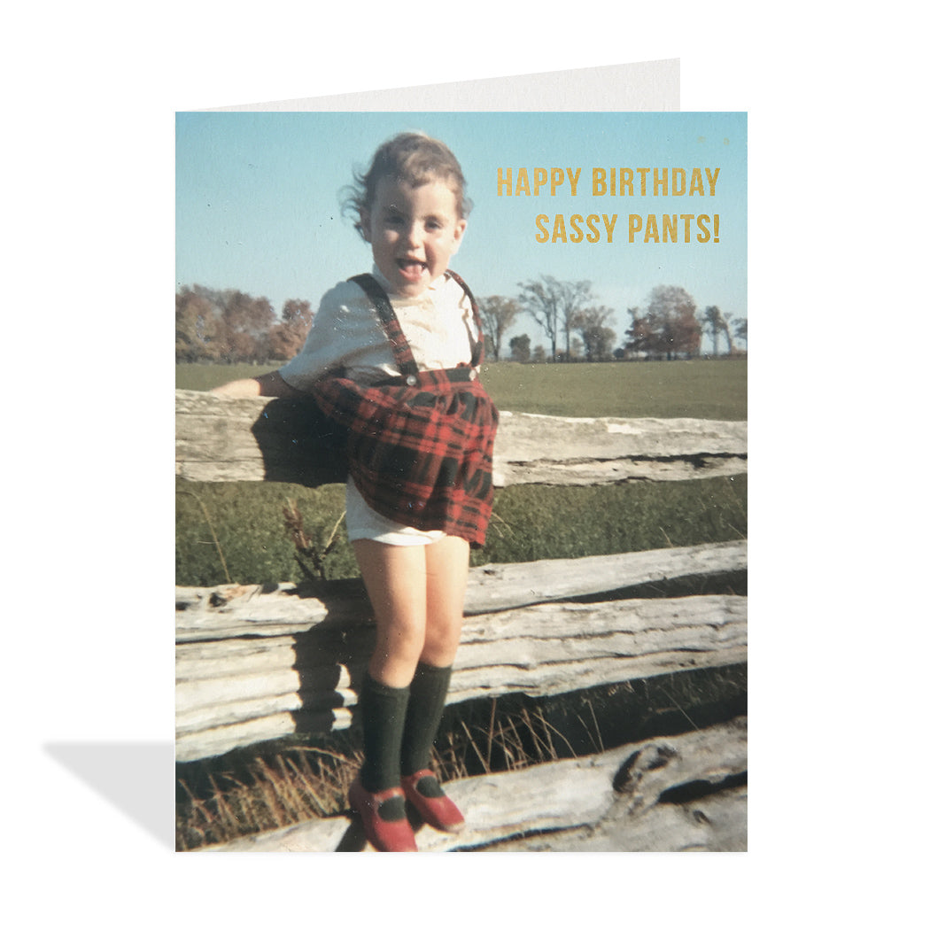 Vintage photograph of a young girl standing on a fence holding down her dress as wind blows her red plaid skirt up. Gold foil typography that reads "Happy birthday sassy pants!"