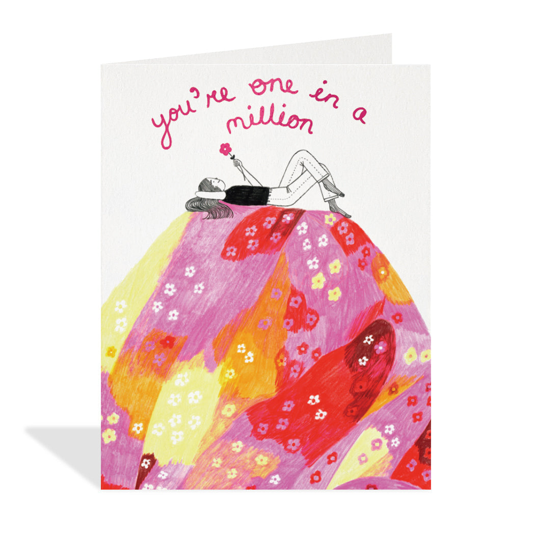 Greeting card design by Chelsea O'Byrne. A cute illustration of a pink hill with a girl laying on top holding a pink foil flower. A sentiment reads "you're one in a million" in pink foil. 