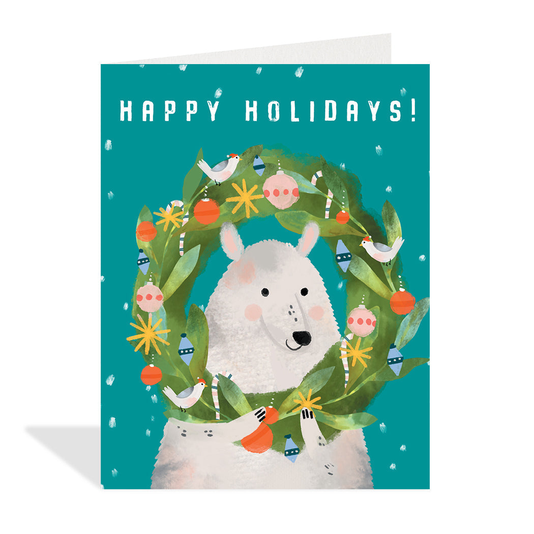 Greeting card illustration by Loose Leaves Paper Goods. Collage-style illustration of a polar bear looking through the middle of a highly decorated wreath, on a teal background with snow falling. Sentiment  eads "Happy holidays"